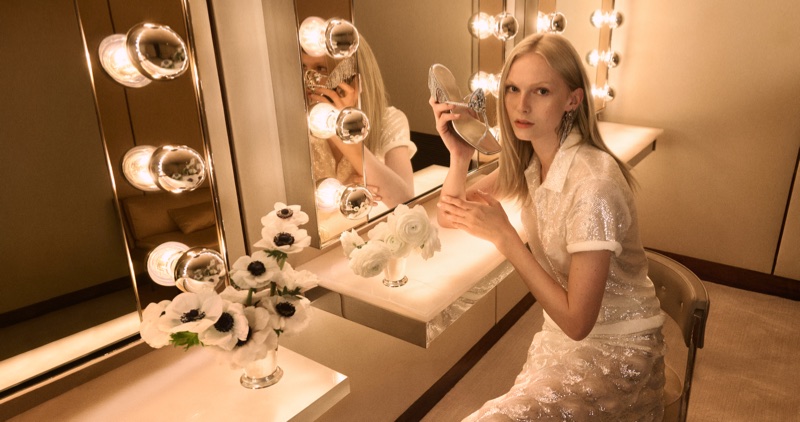 Model Alex Consani seated at an illuminated vanity mirror holding a metallic silver heel, wearing a white sequin top and skirt.