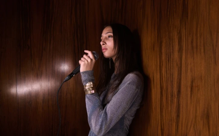 Model holding a microphone against a dark wood-paneled wall, wearing a shimmering silver long-sleeved top and gold watch.