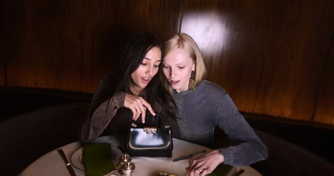 Two female models seated at a restaurant table admiring a black handbag for Tory Burch Holiday 2025