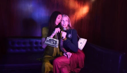 Two female models singing into microphones while seated on a dark leather booth in a dimly lit bar for Tory Burch Holiday 2025.