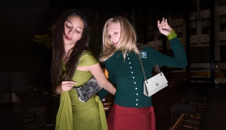 Two female models dancing indoors, one wearing an olive dress and the other a green cardigan with a red skirt