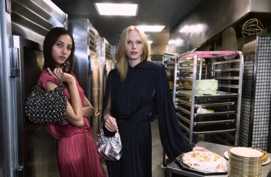 Two models standing in a commercial kitchen holding metallic and embellished handbags for Tory Burch Holiday 2025.