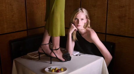 Model seated beside a white-clothed restaurant table with another standing on it, photographed for Tory Burch Holiday 2025.