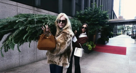 Two models carrying a large pine branch outdoors while wearing sunglasses and winter coats for Tory Burch Holiday 2025