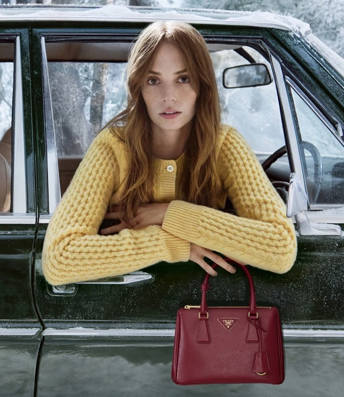 Maya Hawke leaning out of a dark green car window wearing a yellow knit cardigan and holding a red leather handbag for Prada Holiday 2025.