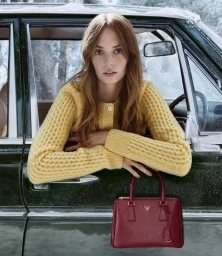 Maya Hawke leaning out of a dark green car window wearing a yellow knit cardigan and holding a red leather handbag for Prada Holiday 2025.
