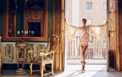 Model standing on a balcony with arms outstretched wearing a patterned mini dress and boots, in an ornate room