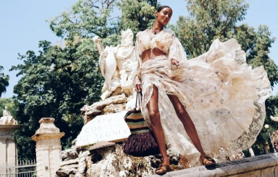 Model in flowing patterned cream two-piece outfit holding a straw fringe bag beside an ornate stone fountain in sunlight.