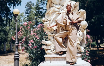 Model in beige utility trousers and matching shirt posing next to a white classical stone statue outdoors.