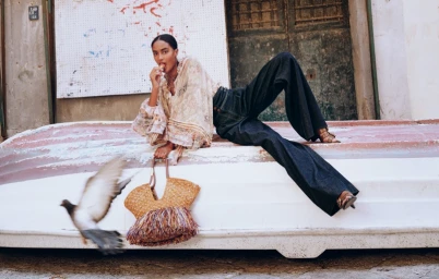 Model lounging on a white boat structure with a straw fringe bag as a pigeon flies past in the foreground.