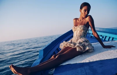 Model Sacha Quenby in a patterned strapless dress seated on a blue boat deck at sea by Julia Noni.