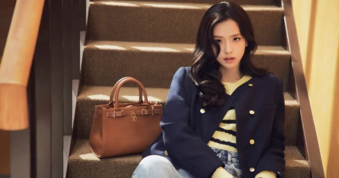 Jisoo sitting on indoor wooden stairs next to a brown leather handbag, wearing a navy jacket and striped sweater for Tommy Hilfiger.