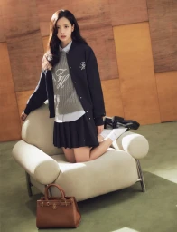 Jisoo kneeling on a cream armchair wearing a dark bomber jacket, grey knit sweater, pleated skirt, and a brown handbag nearby.
