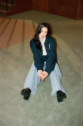 Jisoo seated on a green carpet wearing a dark navy cardigan, white shirt, and wide-leg light blue jeans for Tommy Hilfiger.