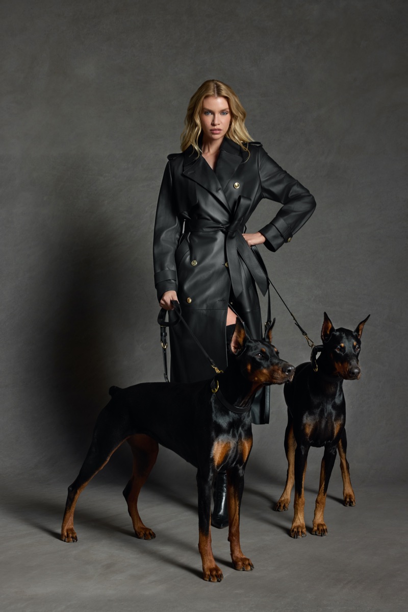 Stella Maxwell in a belted black leather trench coat posing with two Doberman pinschers on leashes in a studio setting.