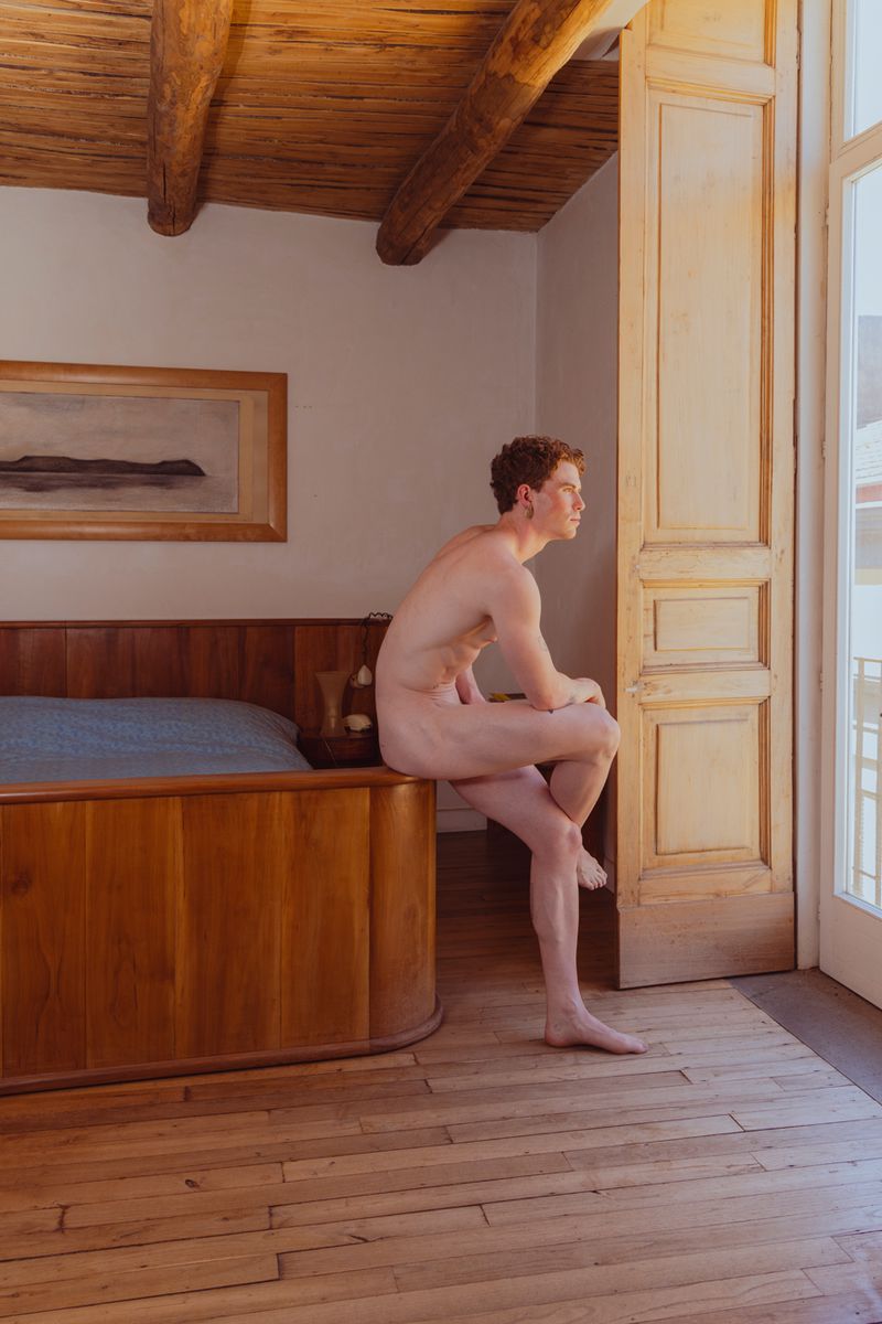 Nude male figure seated on bed edge, gazing through open door in sunlit room; bedroom interior with wood floors and ceiling beams.