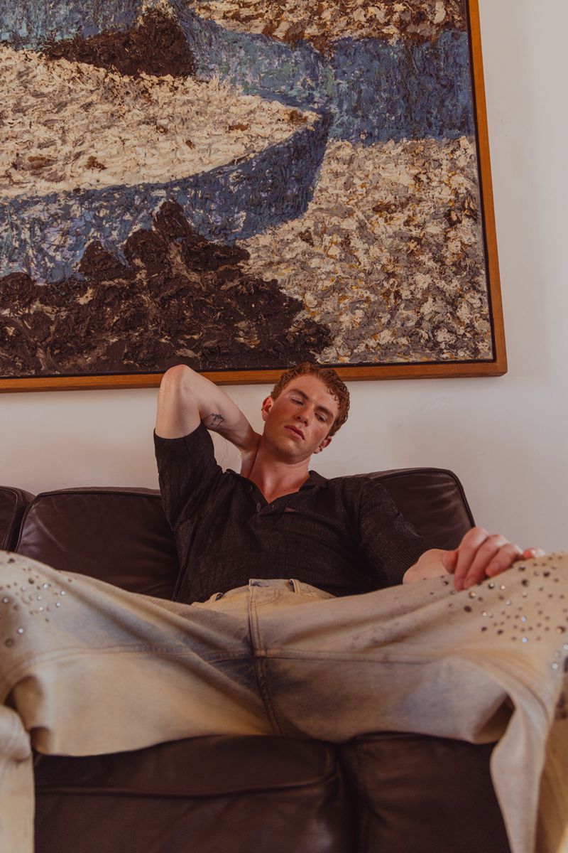 A red-haired man relaxes on a brown leather couch in a black shirt and light wash jeans, in front of a painting in a warmly lit room.