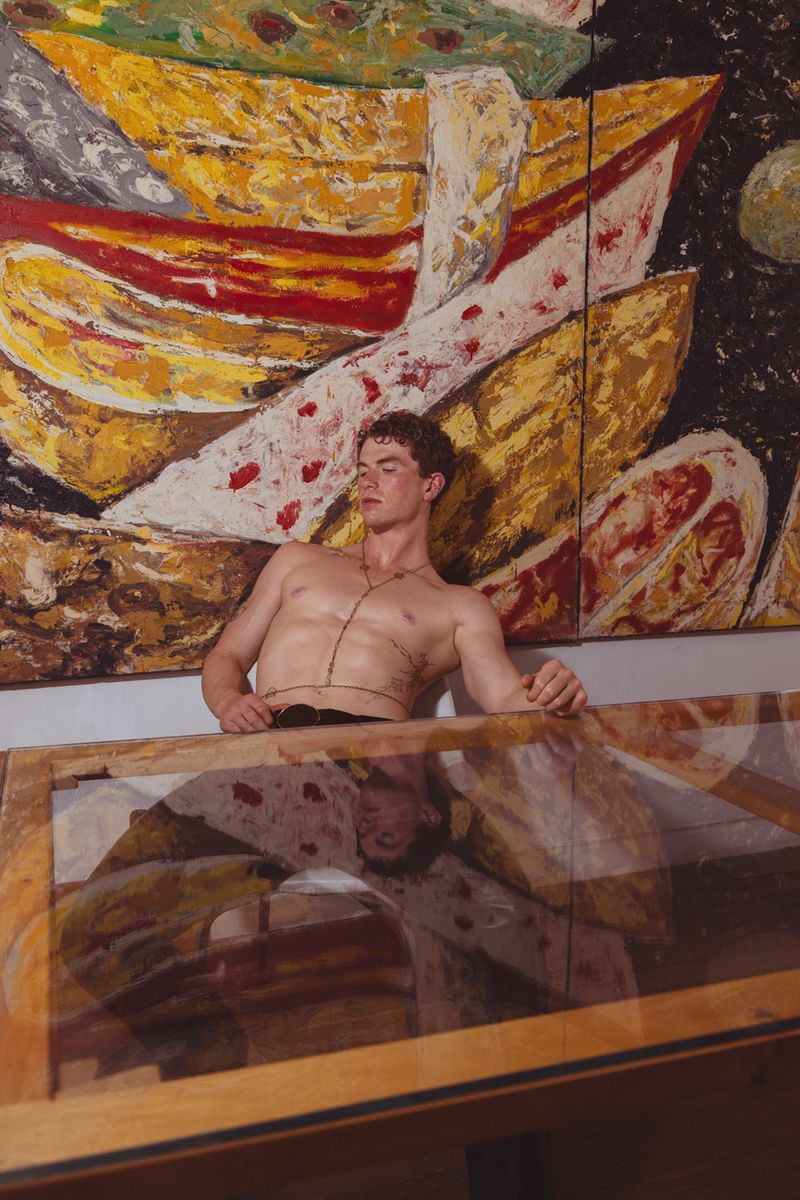 Male torso with necklace against abstract painted artwork, reflected in glass surface, studio lighting, artistic nude male portrait.