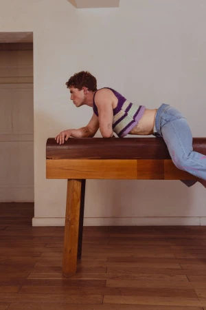 Young adult male poses on vintage gymnastic vault in a light-filled room with a crocheted top and jeans for an athletic fashion editorial.
