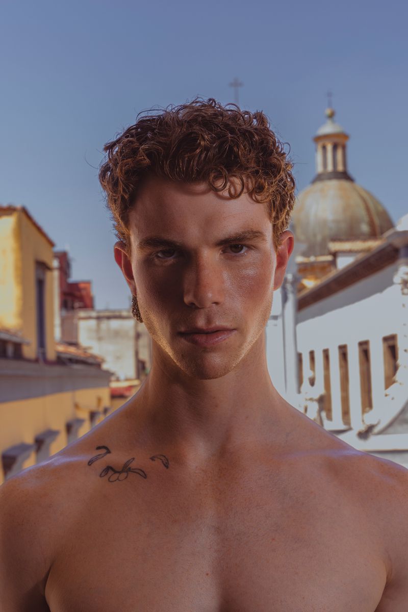 Portrait of a bare-chested man with curly red hair outdoors in front of buildings on a sunny day, with a shallow depth of field.