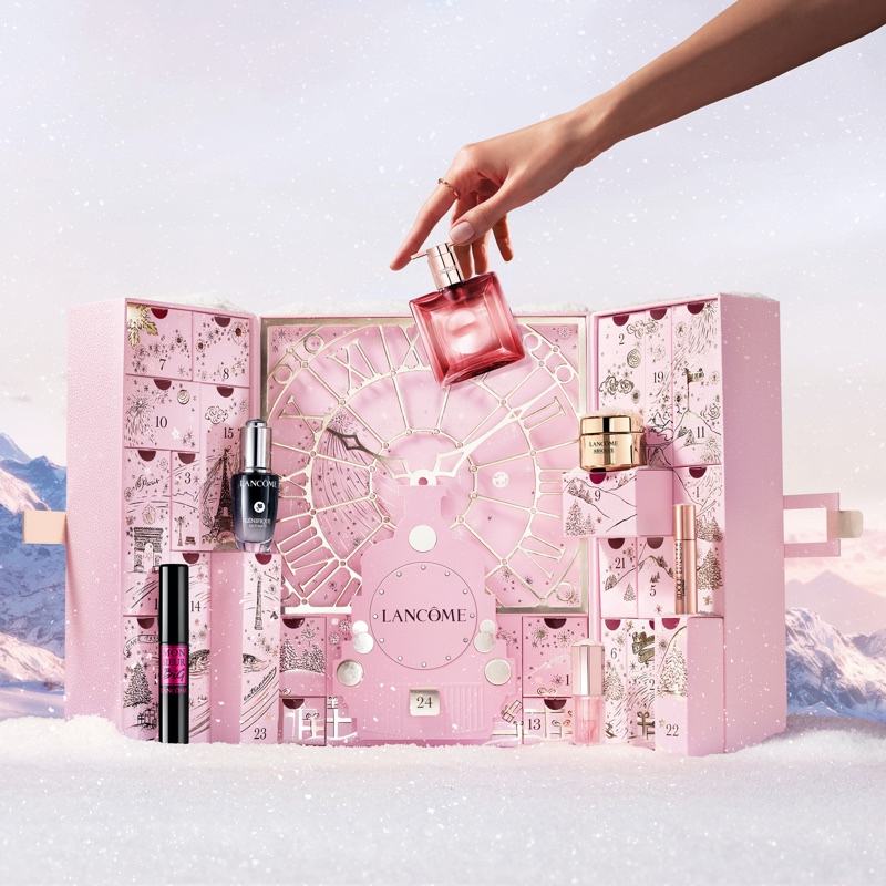 Hand holding a pink perfume bottle above an open pink Lancôme holiday advent calendar in a snowy alpine setting.