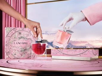 Two hands reaching towards pink perfume bottles on a decorated tabletop with a winter mountain landscape in the background