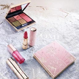 Lancome holiday makeup collection featuring eyeshadow palette, pink lipstick, lip gloss, and festive packaging on a snowy background