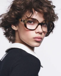 Model with short curly brown hair wearing tortoiseshell glasses and a black top with white collar against white background