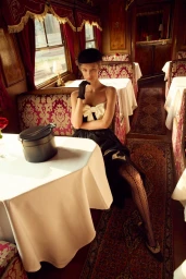 Model Kris Grikaite in a black dress with white bow seated inside a vintage train dining car for For Love & Lemons Winter 2025 campaign.