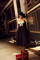 Model Kris Grikaite in a black and white dress posing on red steps beside a train car for For Love & Lemons Winter 2025