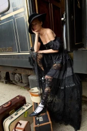 Model Kris Grikaite in a black lace dress and wide-brim hat seated on a vintage train car with old suitcases