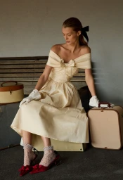 Model Kris Grikaite seated on vintage luggage wearing an off-shoulder cream dress with matching gloves and red heels.