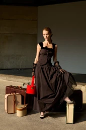 Model Kris Grikaite in a dark dress holding a red vanity case amid vintage luggage for For Love & Lemons Winter 2025 campaign.