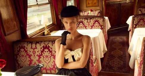 Kris Grikaite in a black hat and dress inside a luxury train dining car for For Love & Lemons Winter 2025 by Camilla Akrans.