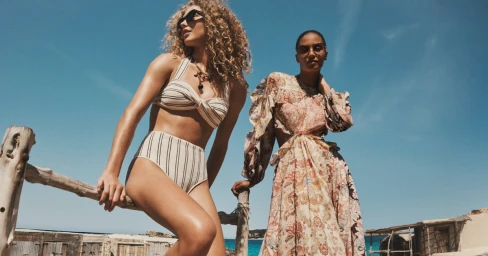 Two models posing outdoors against a clear blue sky, one wearing a striped two-piece swimsuit and the other a floral patterned dress.