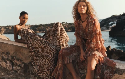 Two models in patterned resort wear posing against a stone wall overlooking water under an open sky.