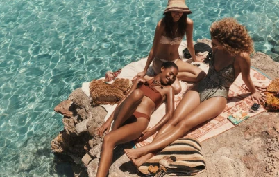Three female models relaxing on a patterned towel spread across rocky coast beside clear blue ocean water