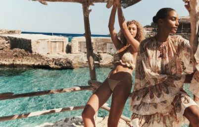 Two models posing near a wooden structure by a rocky coast and turquoise ocean for Zimmermann Resort Swim 2026.