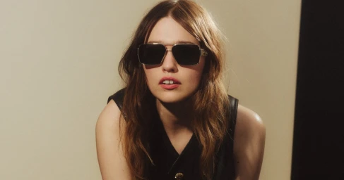 Aimee Lou Wood wearing dark sunglasses and a black top against a minimalist neutral studio background.
