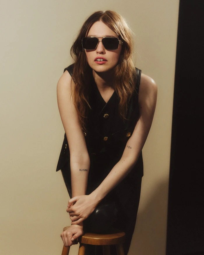 Aimee Lou Wood wearing dark sunglasses and a black vest leaning on a stool for Versace Greca Eyewear.