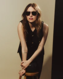 Aimee Lou Wood wearing dark sunglasses and a black vest leaning on a stool for Versace Greca Eyewear.