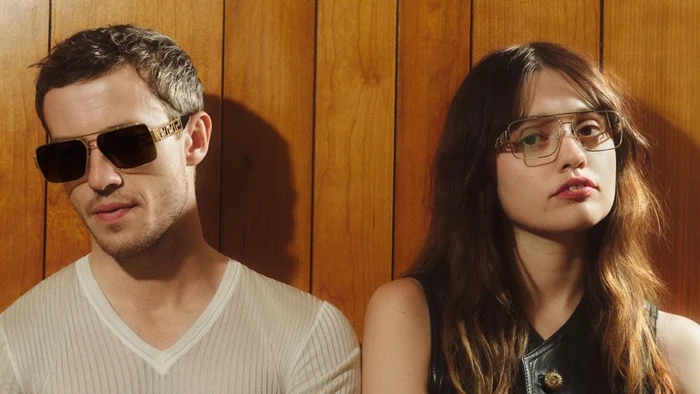 Joseph Quinn and Aimee Lou Wood wearing Versace Greca Eyewear against a wood paneled wall