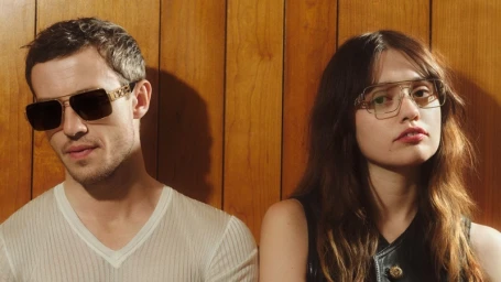 Joseph Quinn and Aimee Lou Wood wearing Versace Greca Eyewear against a wood paneled wall