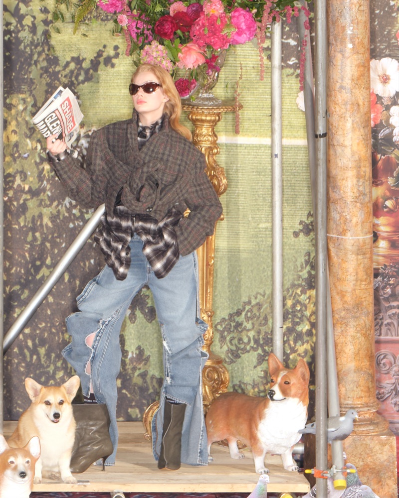 Model in dark sunglasses holding a newspaper and wearing a plaid jacket over distressed denim jeans with corgis in the foreground.