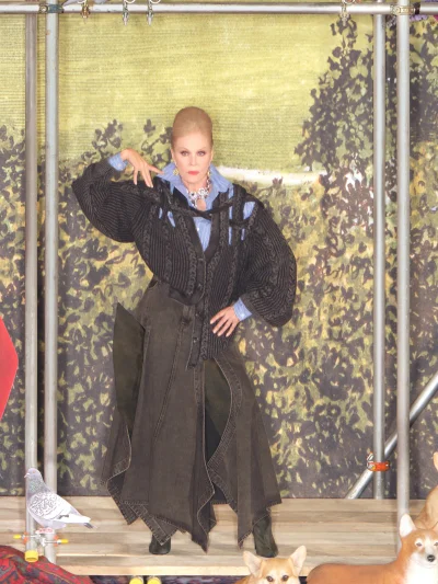 Woman in dark textured knit cardigan and blue shirt posing on a stage with painted backdrop and scaffolding.