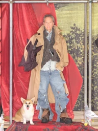 Man wearing beige trench coat and distressed denim jeans standing next to a corgi in a red draped set