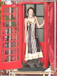 Model wearing a patterned slip dress and tall grey boots posing inside a bright red telephone booth.