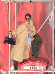 Model in camel coat and dark boots holding a black handbag inside a red telephone box themed set