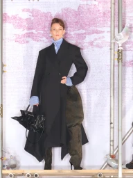 Model in a black overcoat wearing high boots and holding a black handbag against a backdrop with scaffolding and pigeons
