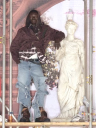 Man wearing a brown hooded jacket and distressed light blue jeans leaning against a classical white statue on scaffolding